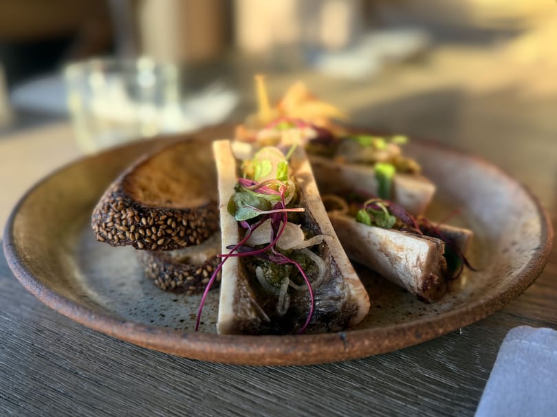 Three bone marrow halves topped with herbs and onions, served with sesame seed toast on a rustic plate.
