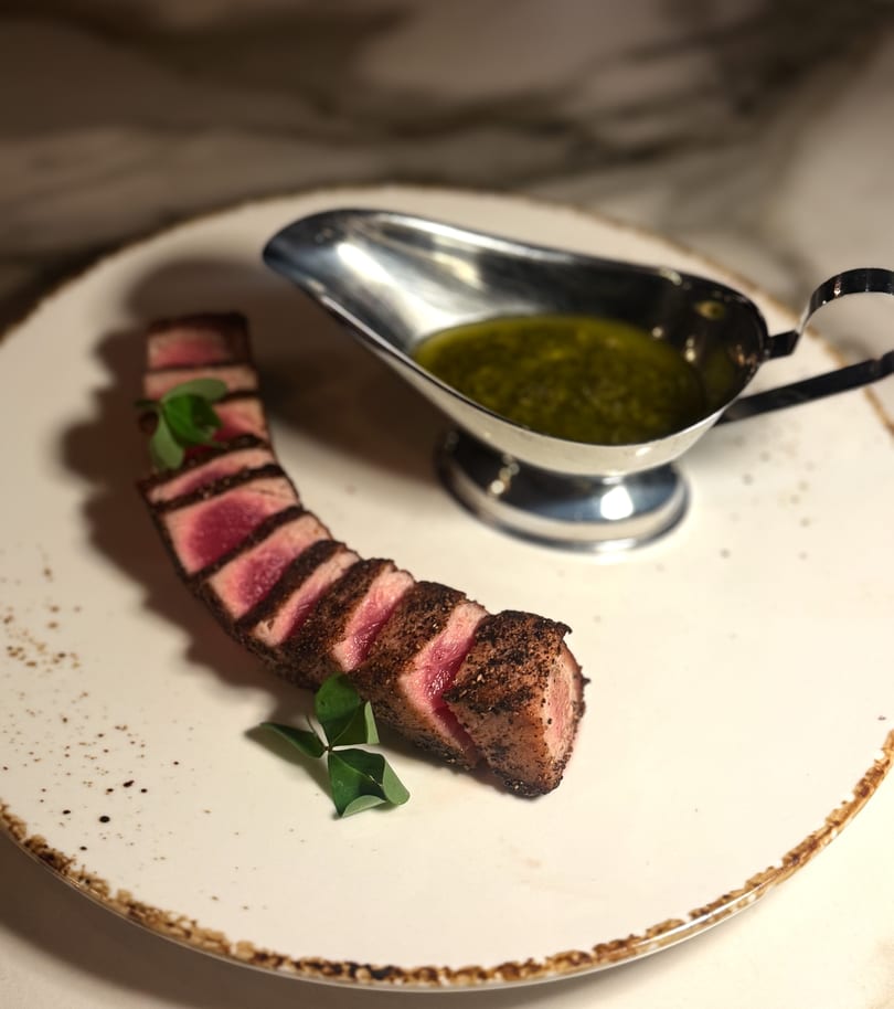 Seared tuna slices with a side of green sauce in a silver gravy boat on a white plate.